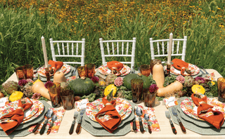 Dining table set for a meal outdoors with floral tablecloth, pumpkins, and autumn-themed decor.